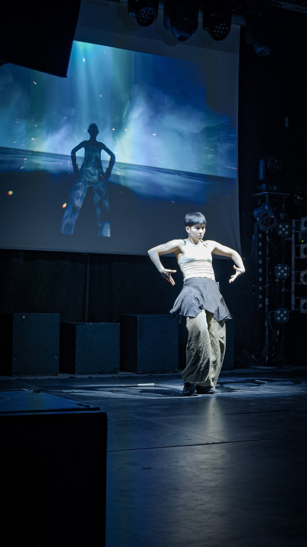 Mathilde on stage, facing the audience, arms out and elbows raised. On the screen behind her, her 3D double holds the same pose above a horizon line.