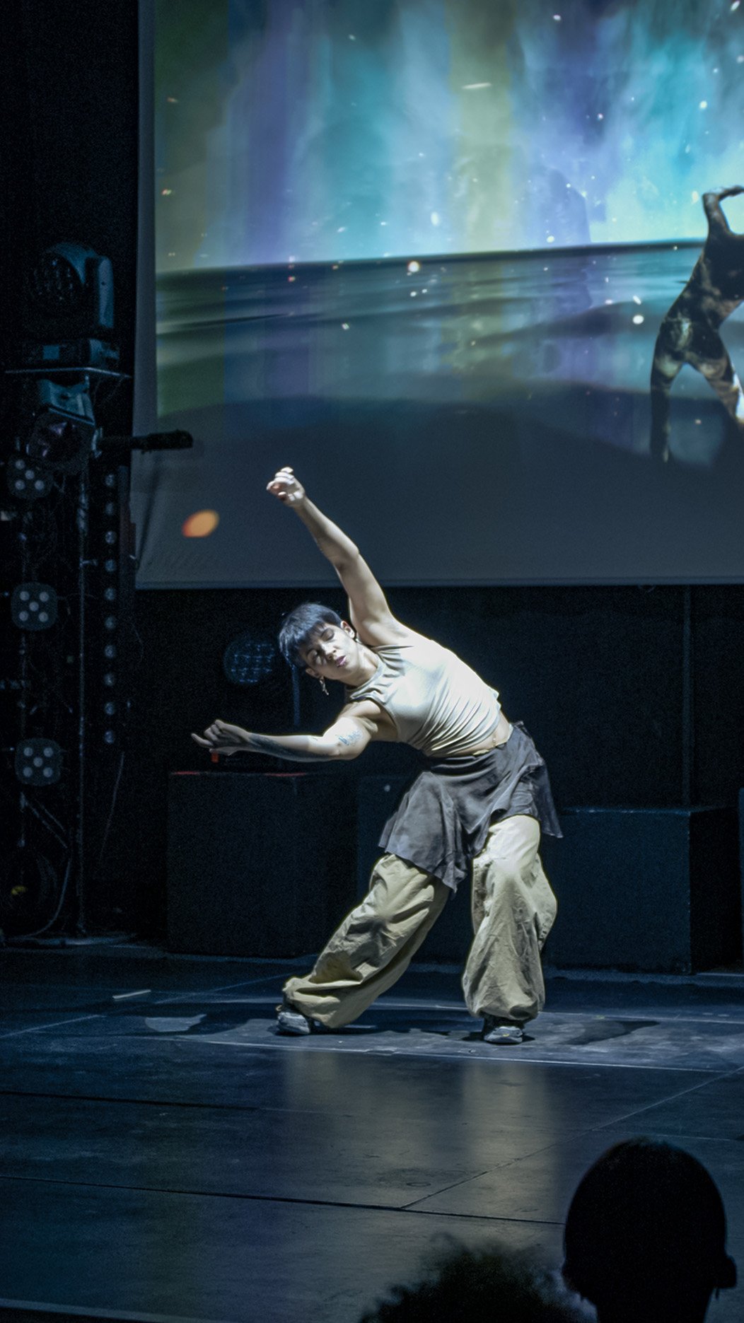 Mathilde leaning sideways, arms spread in opposite directions. Her 3D double appears offset to the right of the screen, among coloured veils.
